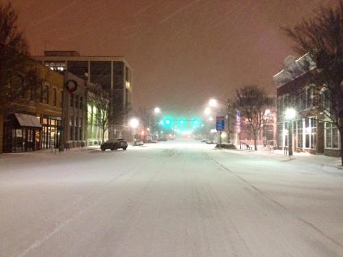 A snowy street at night, with falling snow, empty buildings, and traffic lights glowing in the distance.