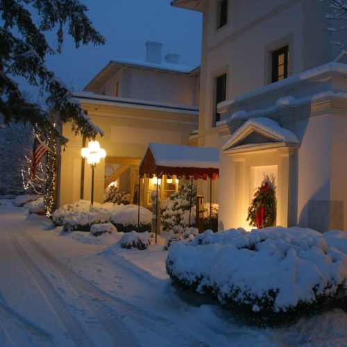 A snowy evening scene of a charming building adorned with lights and a festive wreath, surrounded by snow-covered bushes.