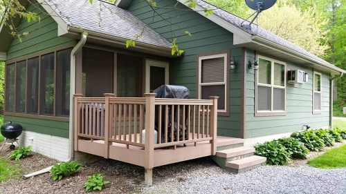 A green house with a wooden deck, grill, and surrounding greenery on a gravel path.