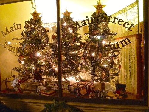 A festive window display featuring two decorated Christmas trees with lights and ornaments at Main Street Emporium.
