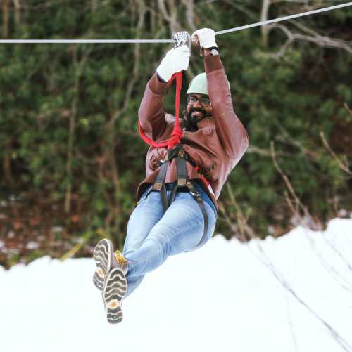 A person in a helmet and harness zip-lining over a snowy landscape, smiling as they glide through the air.