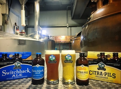 Bottles and glasses of Switchback Ale and Citra-Pils in a brewery setting with brewing equipment in the background.