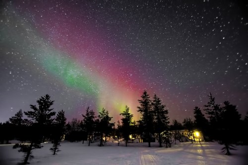 A vibrant aurora borealis lights up the night sky over a snowy landscape, with silhouetted trees and a warm cabin glow.