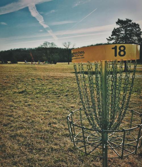 A disc golf basket marked "18" in a grassy field under a clear blue sky with wispy clouds.