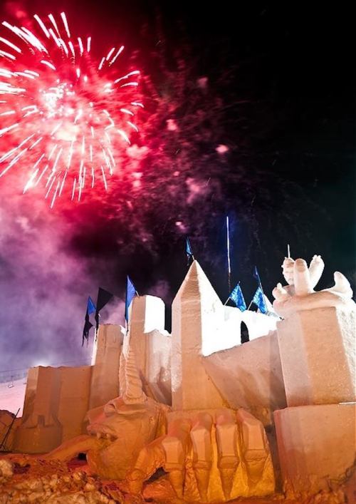 A snow castle illuminated by fireworks, featuring sculptures and blue flags against a night sky.