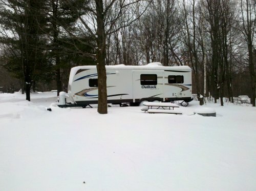 A white RV parked in a snowy landscape, surrounded by trees and a picnic table partially covered in snow.