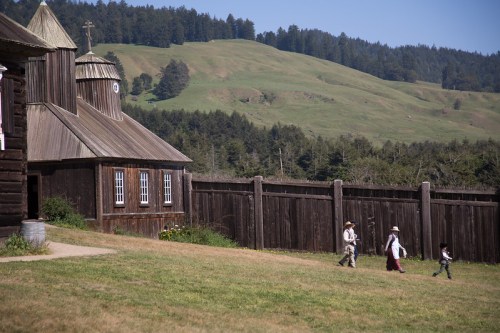 Two people walk along a grassy path near a wooden building, with rolling hills and trees in the background.