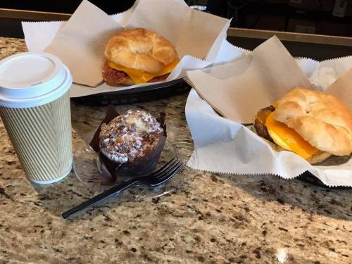A coffee cup, two croissant sandwiches with cheese, and a chocolate muffin on a granite countertop.