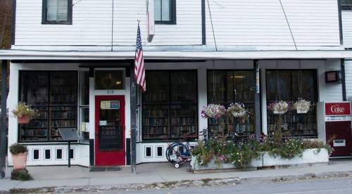 A quaint bookstore with large windows, an American flag, and flower boxes, featuring a bike and a Coke machine outside.