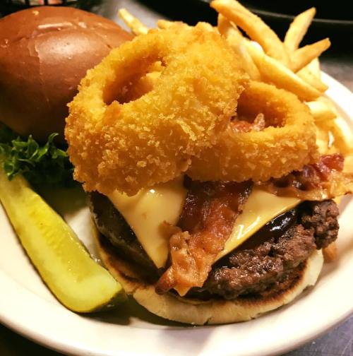 A juicy cheeseburger topped with bacon and onion rings, served with fries, lettuce, and a pickle on the side.