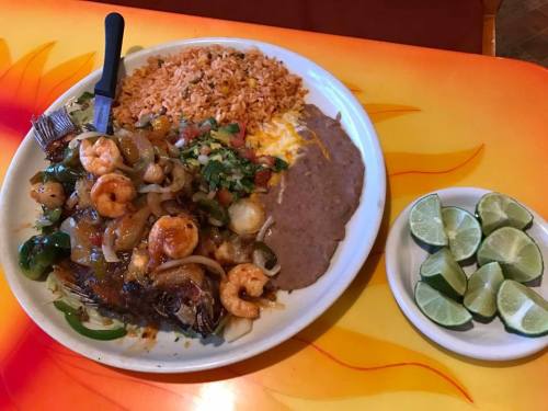 A plate of grilled fish topped with shrimp, onions, and peppers, served with rice, refried beans, and lime wedges.