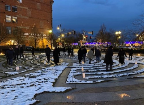 People walking through a snow-covered labyrinth at dusk, with festive lights in the background.