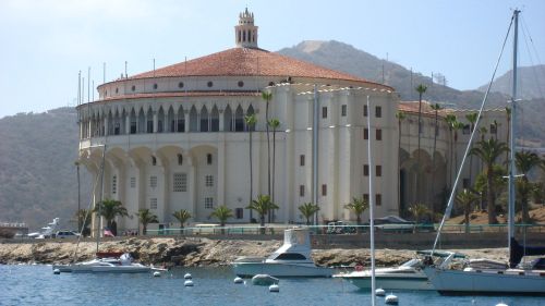 Historic building with a round roof, surrounded by palm trees and boats, set against a mountainous backdrop.
