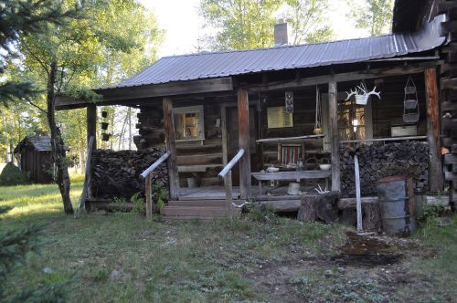 A rustic wooden cabin surrounded by trees, featuring a porch, stacked firewood, and a small outbuilding in the background.
