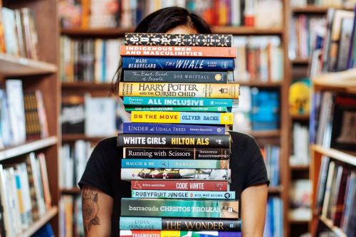 A person stands behind a stack of colorful books in a cozy bookstore, partially obscured by the books.