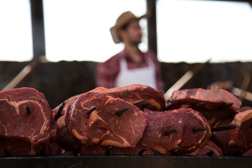 A close-up of raw meat on skewers, with a blurred figure in a hat and apron in the background at a market.