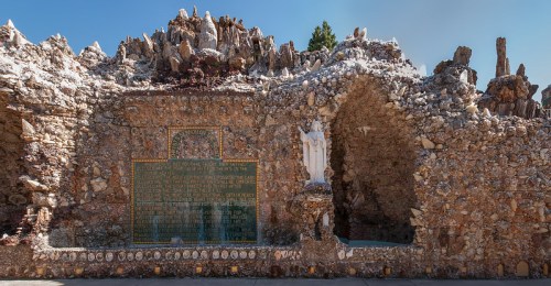 A stone structure with a statue and a plaque, surrounded by rocky formations and greenery under a clear blue sky.