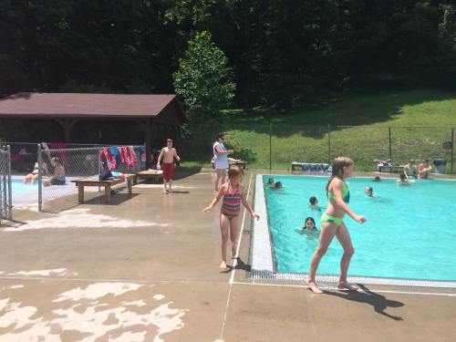 A sunny day at a pool with children swimming, playing, and relaxing on the deck. Trees in the background.