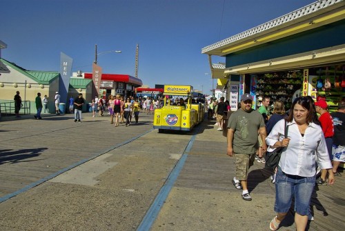 A busy boardwalk scene with people walking, shops on either side, and a yellow sightseeing train in the center.