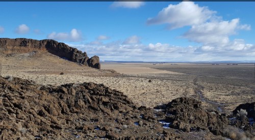 A vast, open landscape with rocky formations, sparse vegetation, and a clear blue sky with scattered clouds.