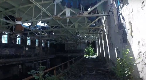 An abandoned industrial building with exposed metal beams, broken windows, and overgrown vegetation inside.