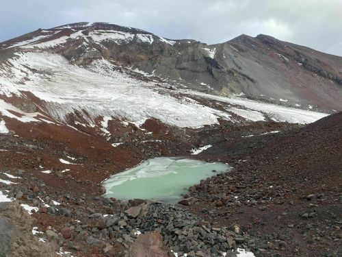 A rocky landscape with a small, icy green lake surrounded by snow-capped mountains and rugged terrain.