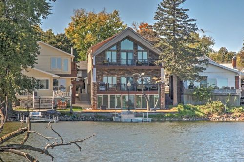 A modern lakeside house with large windows, surrounded by trees and other homes, reflecting on the water.