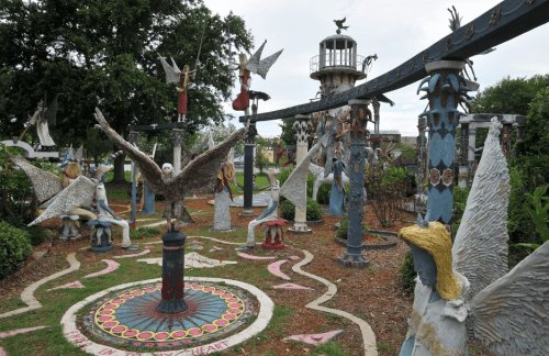 A whimsical garden filled with colorful sculptures of birds and figures, surrounded by greenery and a lighthouse in the background.