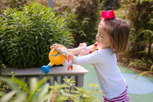 A young girl with a pink bow waters plants in a garden, smiling as she uses a colorful watering can.