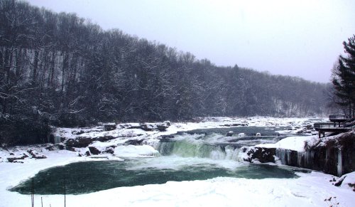 A snowy landscape featuring a waterfall surrounded by icy rocks and a forest of trees in the background.