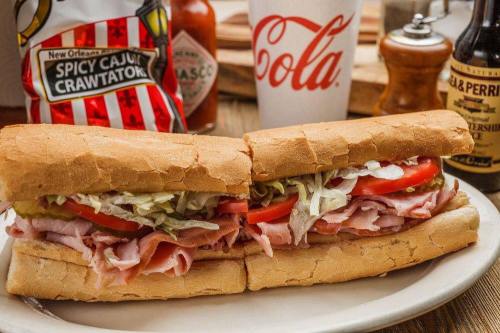 A hearty sandwich with ham, lettuce, and tomato, served with chips and a drink on a wooden table.