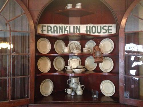 A wooden display cabinet featuring various white dishes and a lantern, labeled "Franklin House" at the top.