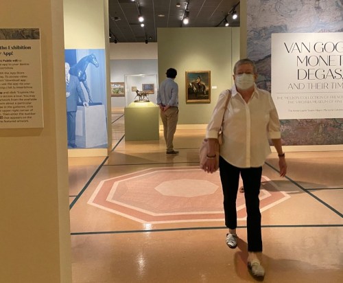 A woman in a white shirt walks through an art gallery, with paintings and sculptures displayed in the background.
