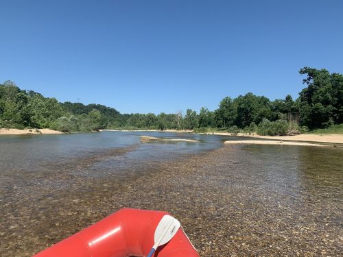 A clear river flows through a lush green landscape, with a red inflatable raft in the foreground under a blue sky.