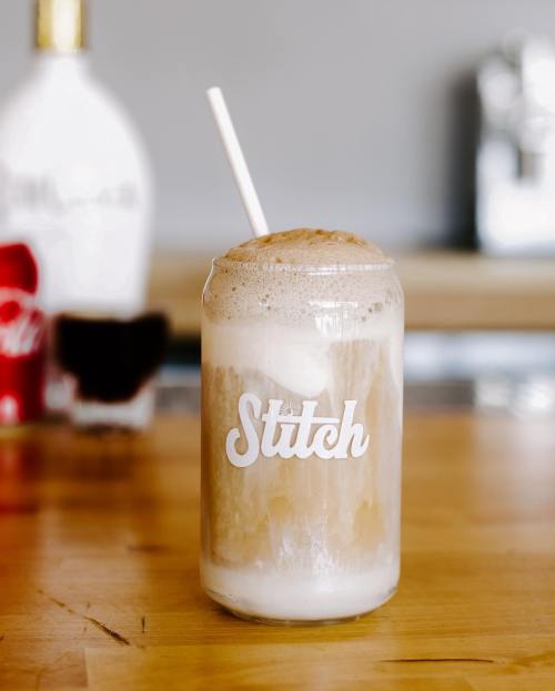 A frothy drink in a glass with a straw, featuring the word "Stitch" on it, set against a blurred background.