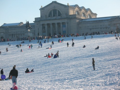 A snowy hill with people sledding, in front of a large historic building on a clear day.