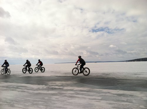 Five cyclists ride on a snowy landscape under a cloudy sky, with a frozen lake stretching out in the background.