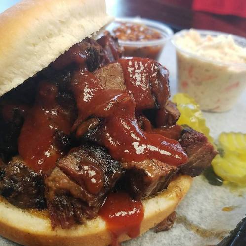 A close-up of a barbecue sandwich with beef, topped with sauce, alongside sides of baked beans and coleslaw.