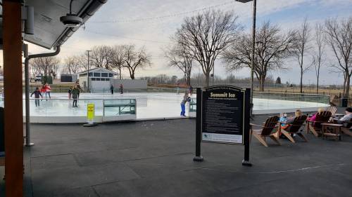 Outdoor ice skating rink with people skating, surrounded by trees and a sign for "Summit Ice." Benches in the foreground.