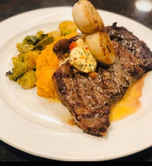 A plated steak with grilled vegetables, creamy squash, and topped with sautéed onions and a dollop of sauce.