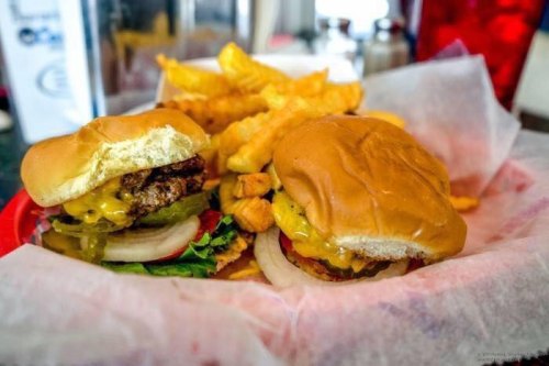 Two cheeseburgers with lettuce, tomato, and pickles, served with crinkle-cut fries in a red basket.