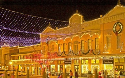 A festive street scene at night, adorned with colorful holiday lights and decorations on buildings and trees.