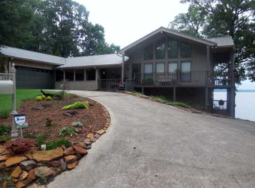 A modern lakeside house with a sloped driveway, large windows, and a landscaped yard. Trees line the background.