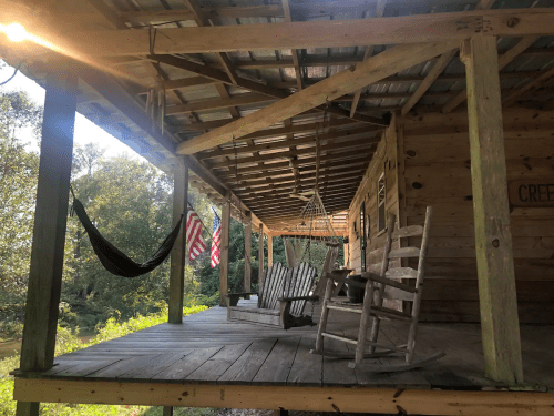 A rustic porch with rocking chairs and a hammock, surrounded by trees and featuring American flags.