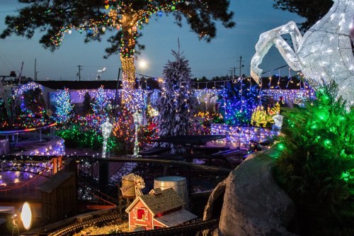A festive scene with colorful holiday lights illuminating trees, decorations, and a miniature train setup at dusk.