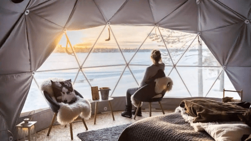 A person sits in a geodesic dome, gazing at a snowy landscape and sunset through large windows. Cozy interior decor.