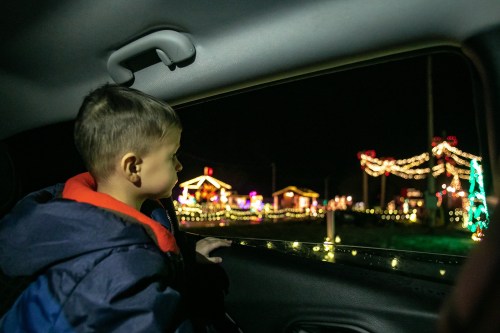 A child gazes out of a car window at colorful holiday lights decorating nearby houses at night.