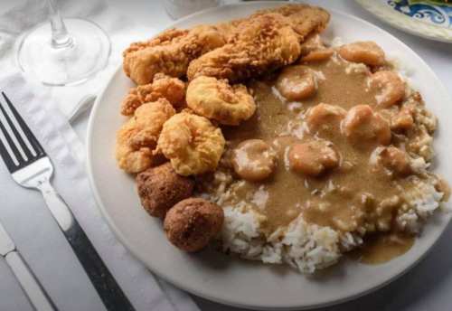 A plate of fried fish, shrimp in gravy, and rice, accompanied by hushpuppies, set on a table with utensils.