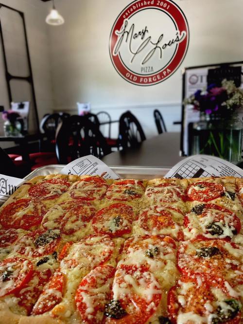 A close-up of a tray of pizza topped with pepperoni, cheese, and fresh tomatoes, with a restaurant interior in the background.