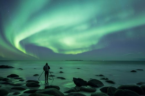 A person stands on rocky shore, capturing the vibrant green aurora borealis lighting up the night sky.